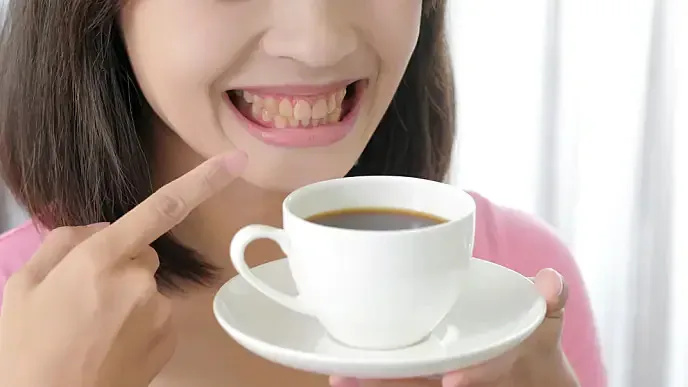 Smiling woman pointing at her teeth while holding a cup of coffee.